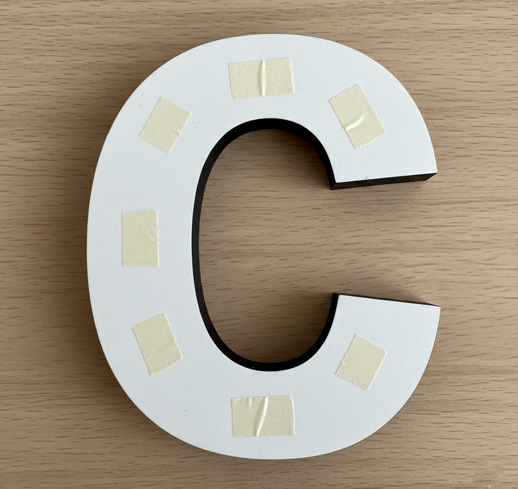 テーブルの上に裏返しに置かれたカルプ文字の裏面に、外周と中央に細く両面テープを貼っている手元アップの画像