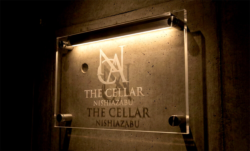 西麻布のコンクリート打ちっぱなしの壁に設置された、透明で厚みのあるアクリル看板。高級感のあるロゴが浮かび上がる